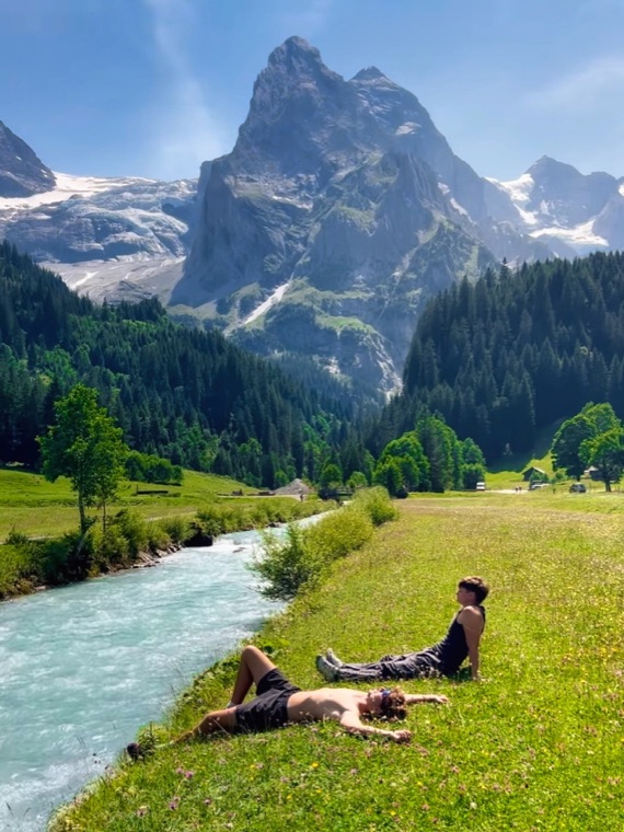 Photo of a person resting on the grass by a turquoise river below a Swiss peak.